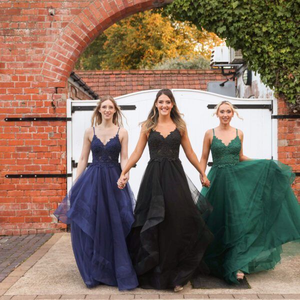 Three women in formal gowns holding hands, walking near a brick archway.