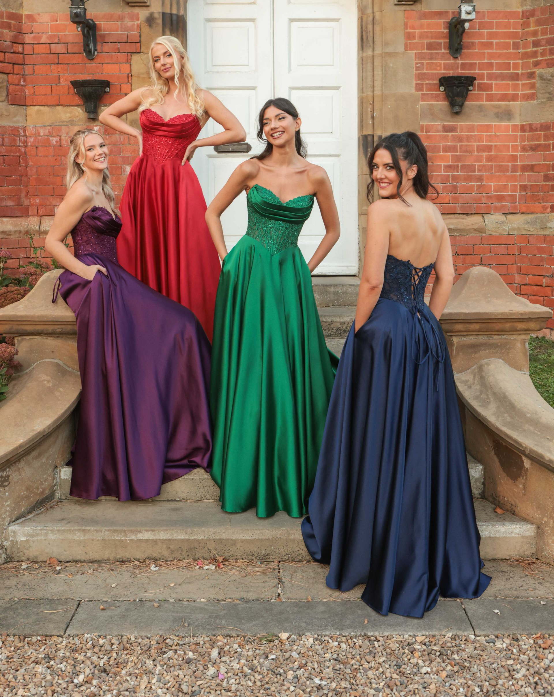 Four women in strapless formal gowns posing on steps of a brick building. Dresses are purple, red, green, and blue.
