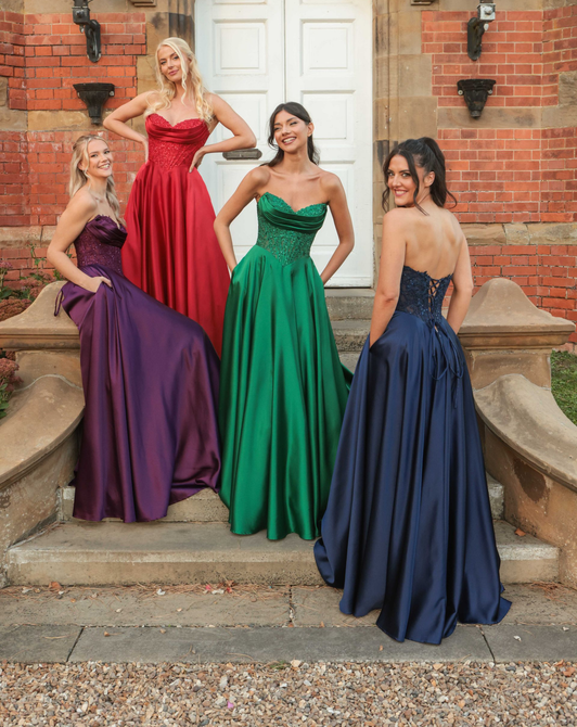 Four women in strapless formal gowns posing on steps of a brick building. Dresses are purple, red, green, and blue.