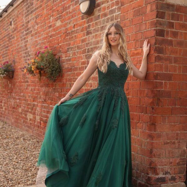 Woman in green formal dress leans against a brick wall, smiling.