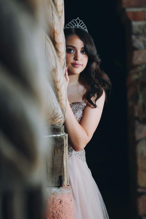 Young person in formal dress and tiara, posing next to a textured pillar, looking at the camera.