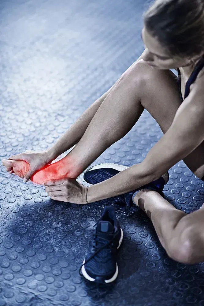 Woman holding foot, highlighting red pain spot; sitting on blue gym floor next to a shoe.