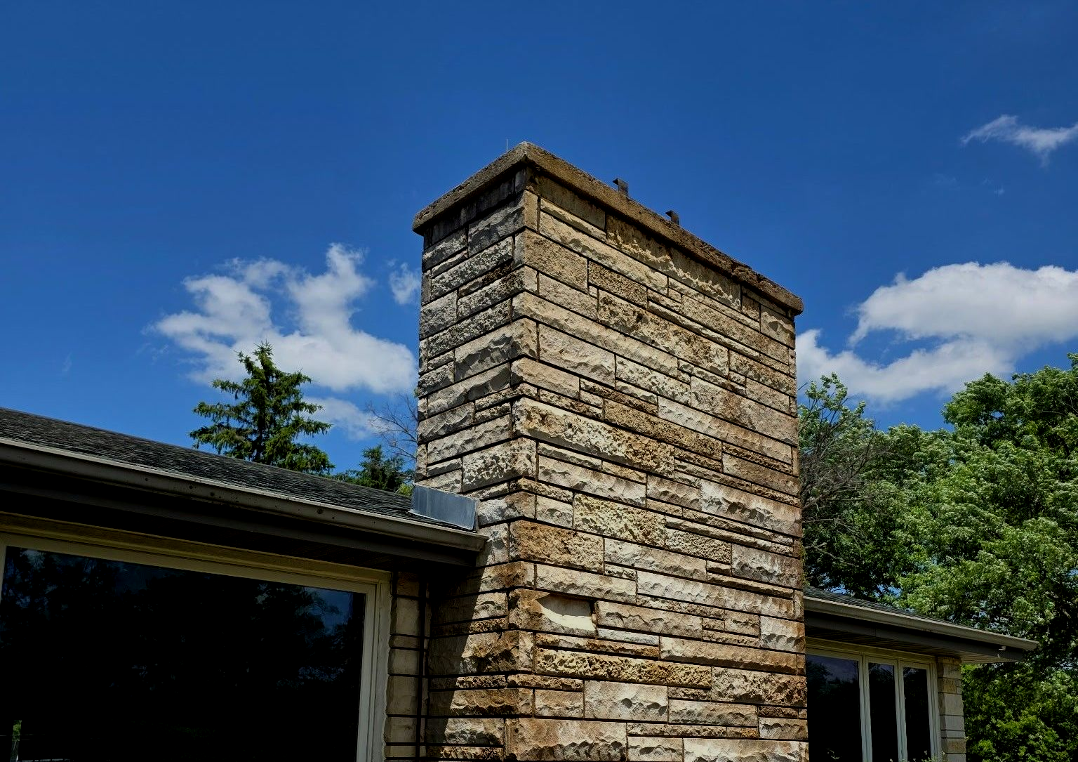 Stone chimney against a bright blue sky, part of a house roof visible below.