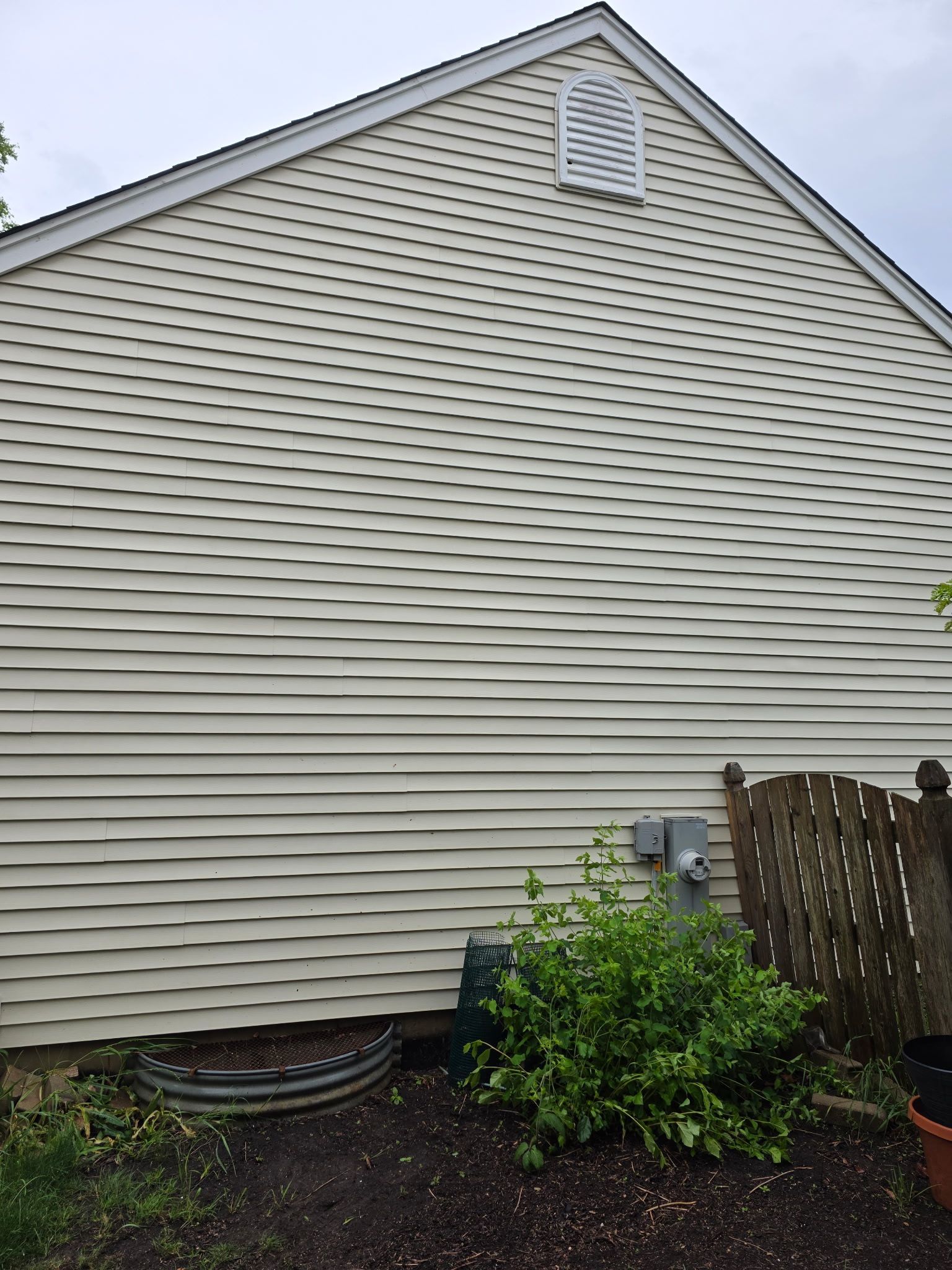 Beige siding on a house with wavy distortions, a small vent, and a garden.
