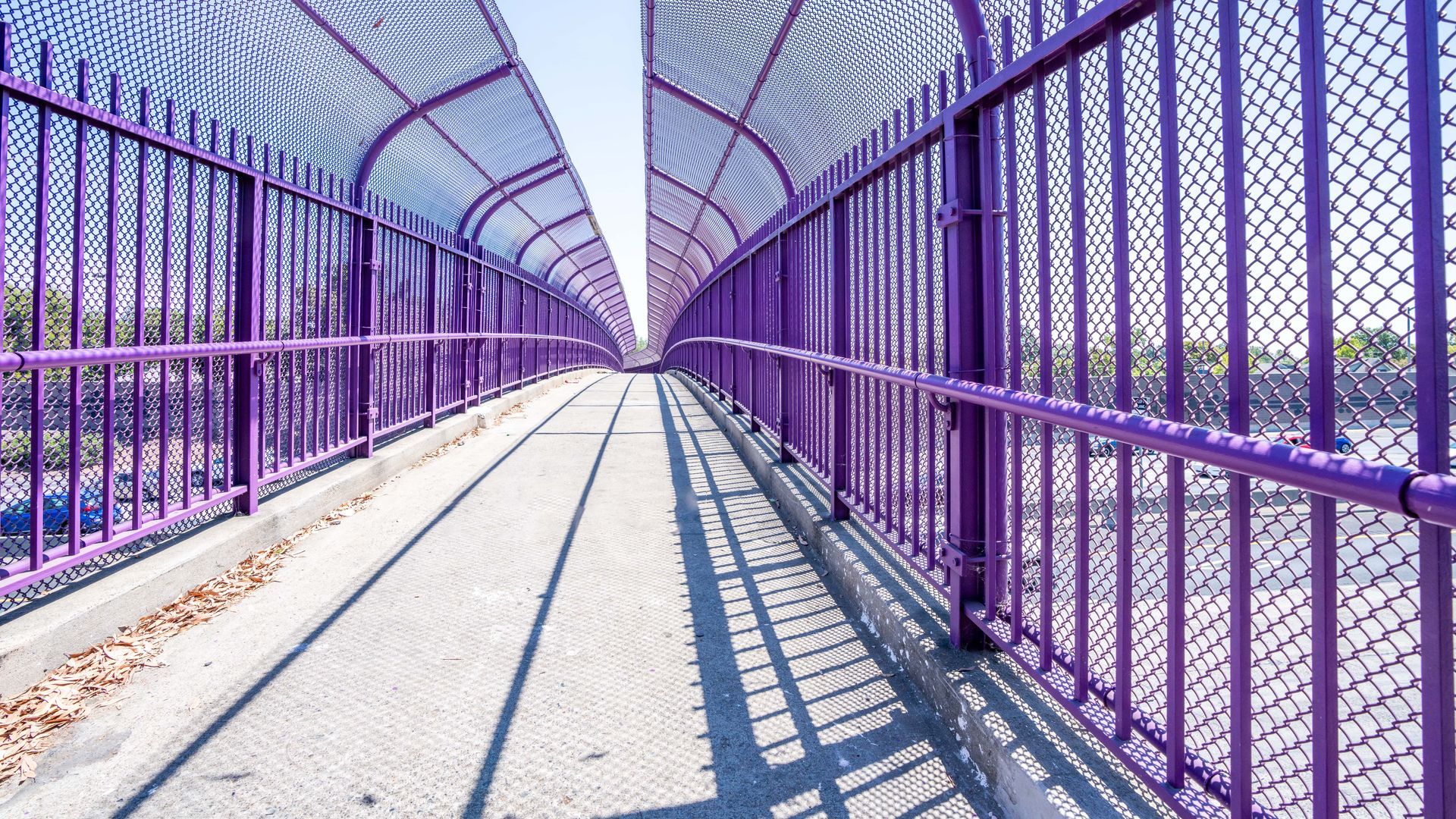Purple pedestrian bridge with metal railings and chain link fencing, casting long shadows.