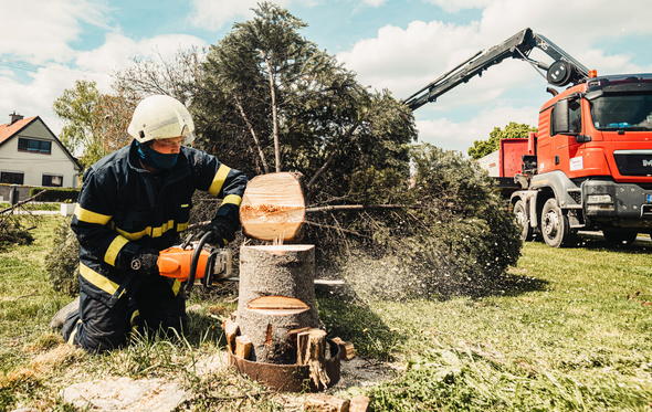 Firefighter using a chainsaw to cut a tree trunk on a lawn, crane truck in background.