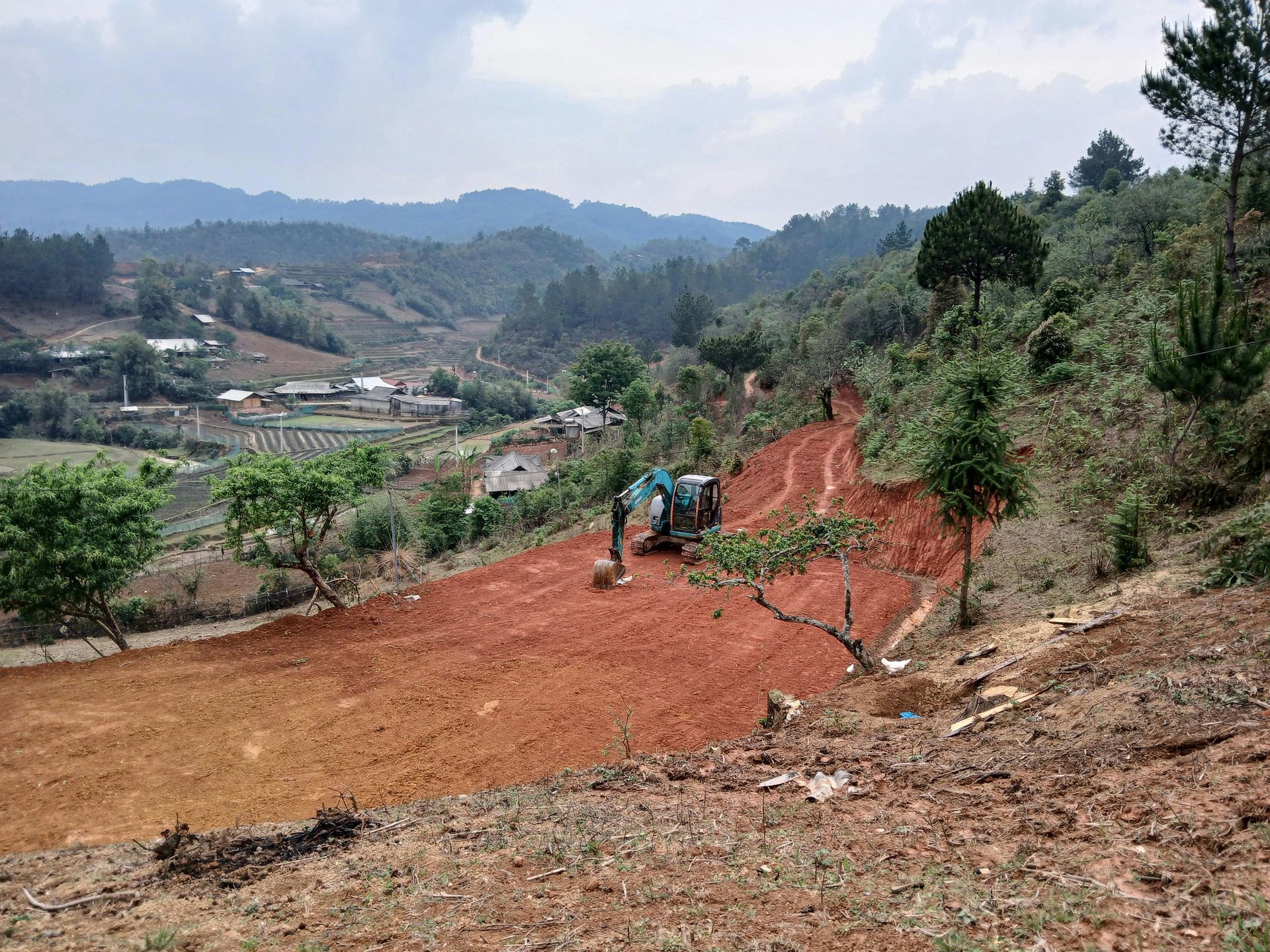 Terraced hillside with red soil road, workers, and a rural valley under a cloudy sky