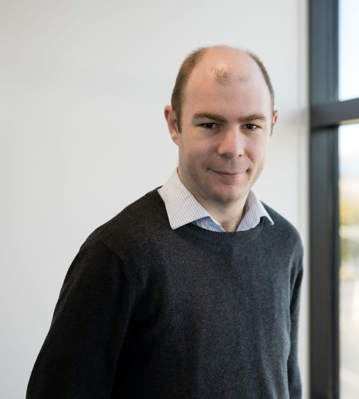 A bald man wearing a black sweater and a white shirt is standing in front of a window.