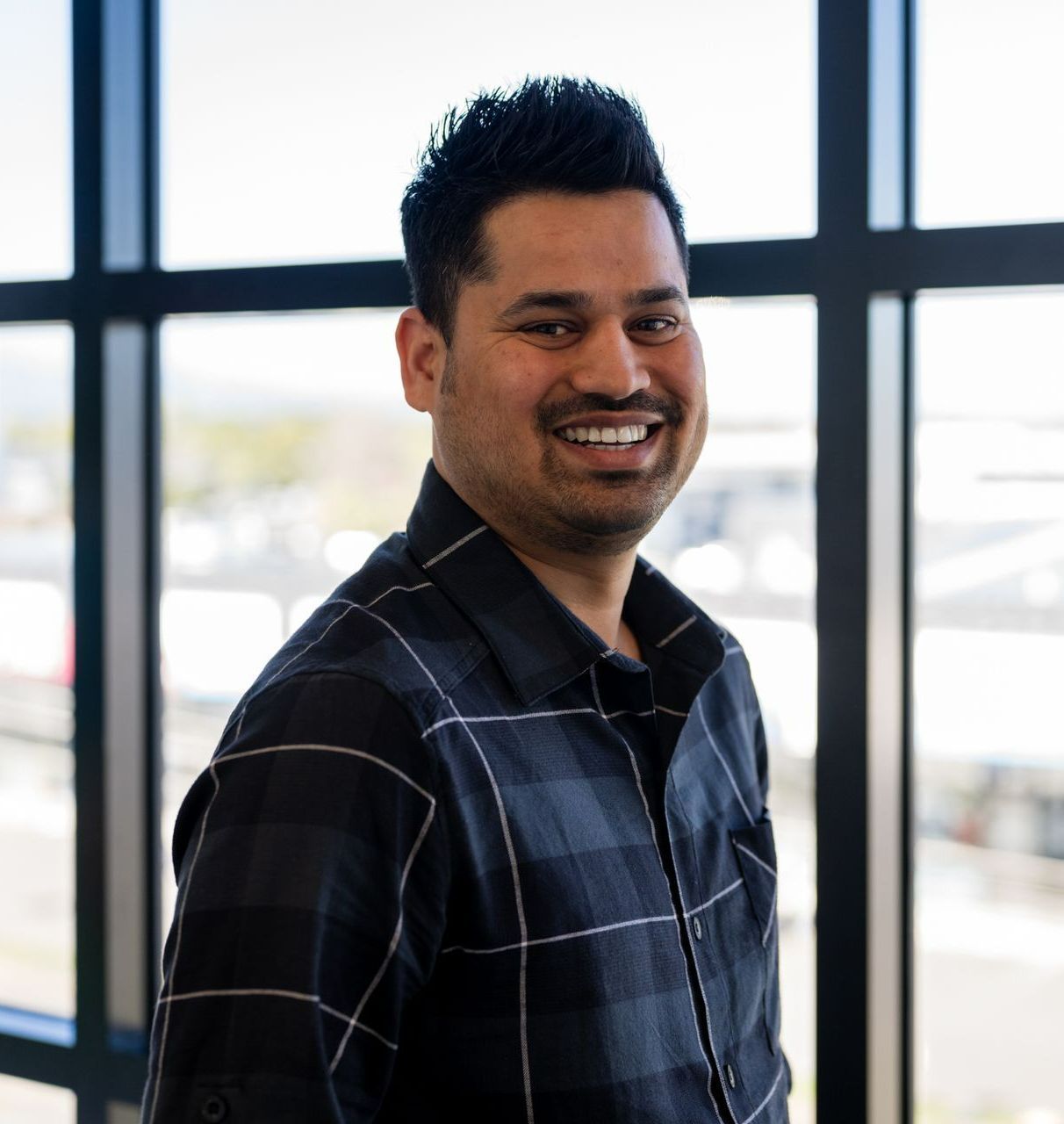 Manish Rawat in a plaid shirt is smiling in front of a window