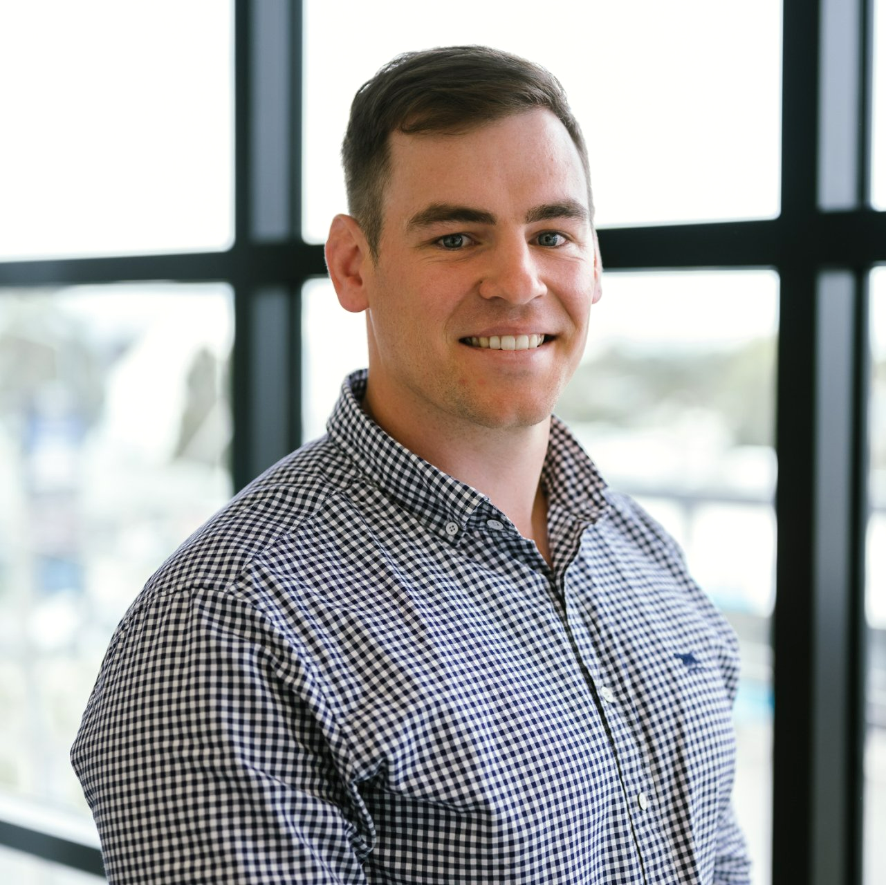 A man in a plaid shirt is smiling in front of a window.