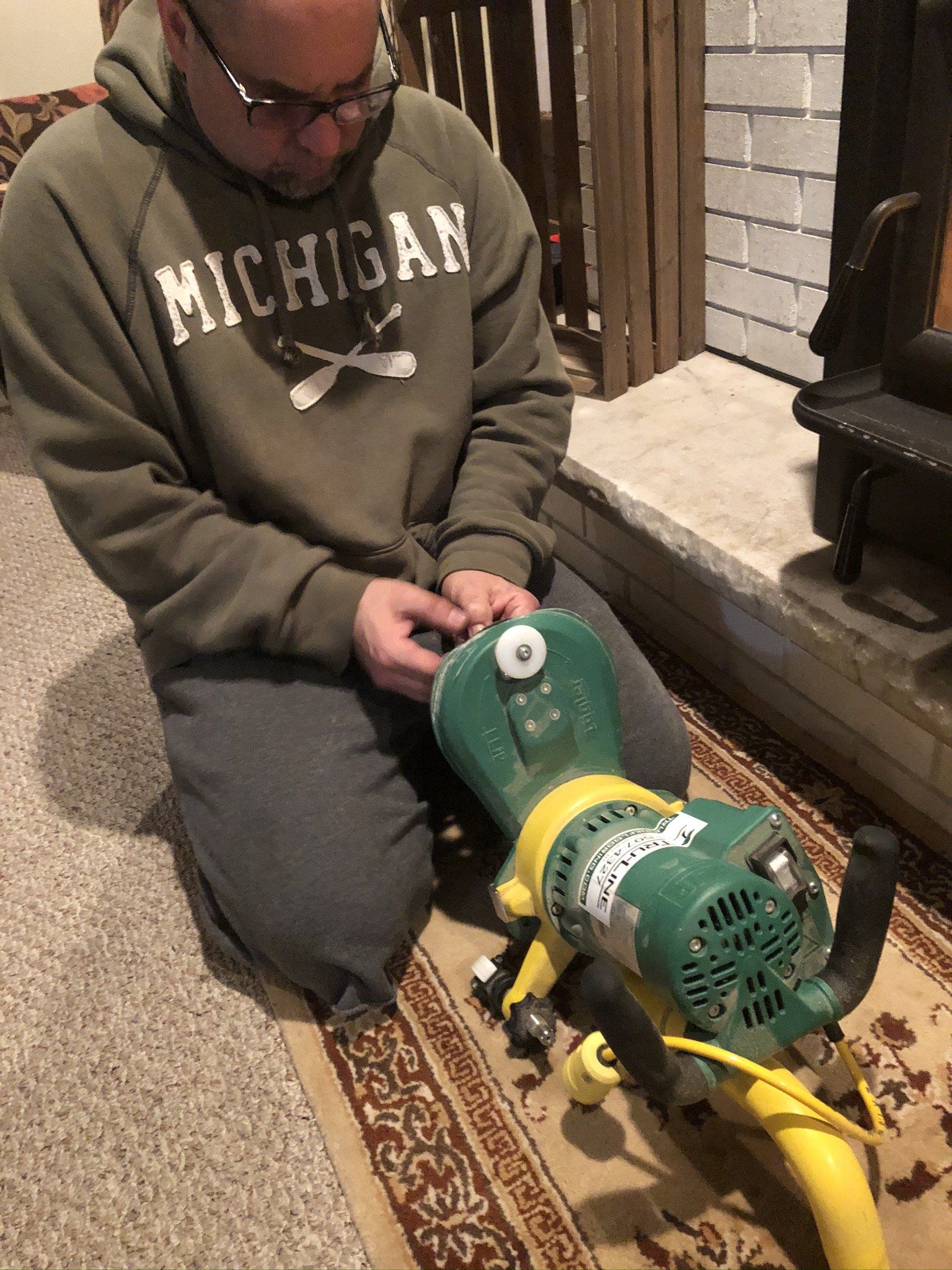 A man in a michigan sweatshirt is kneeling down next to a green and yellow machine.