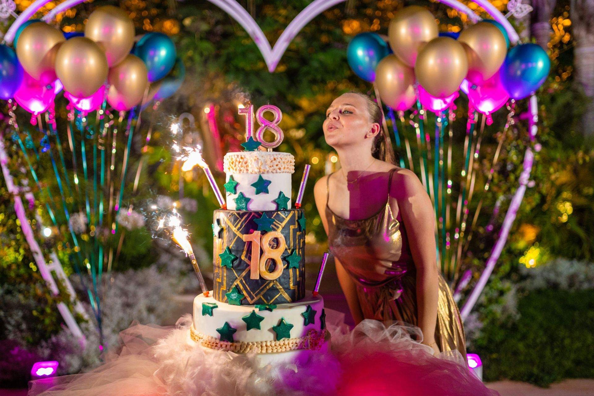 Birthday cake with gold 18 topper, balloons, sparklers, and a woman in a magenta dress at an outdoor celebration