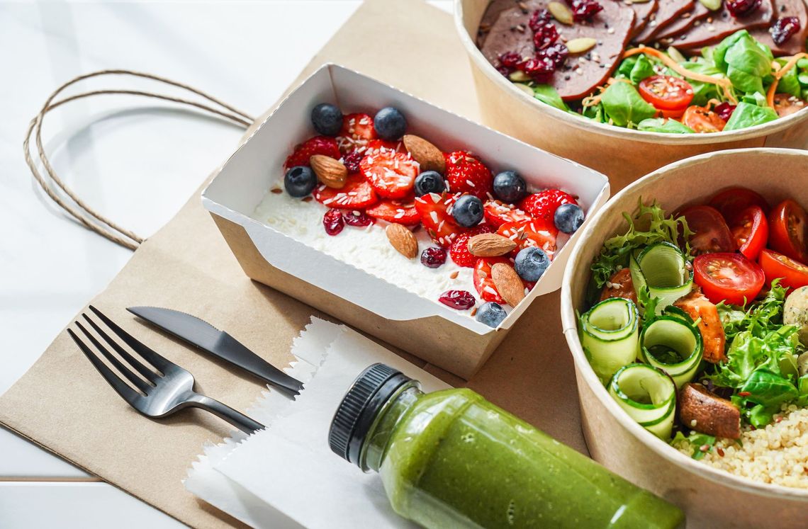 Takeout containers with salad, yogurt, and a green juice on a paper bag with cutler
