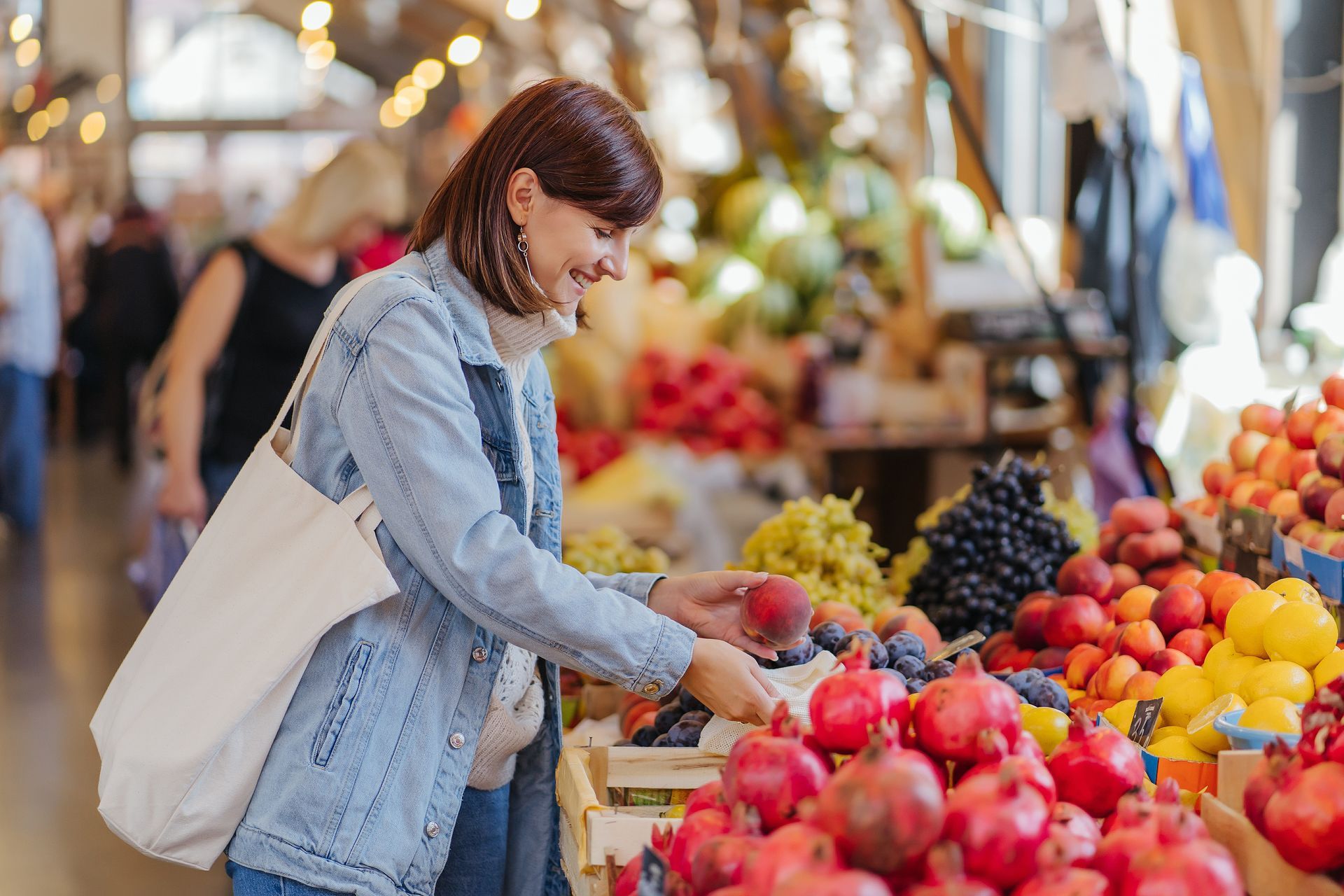 Woman in denim jacket selecting fruit at a market, holding a tote bag.