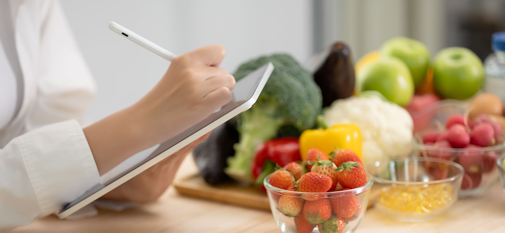 Person writing on a tablet with a stylus, fruits and vegetables in the background.
