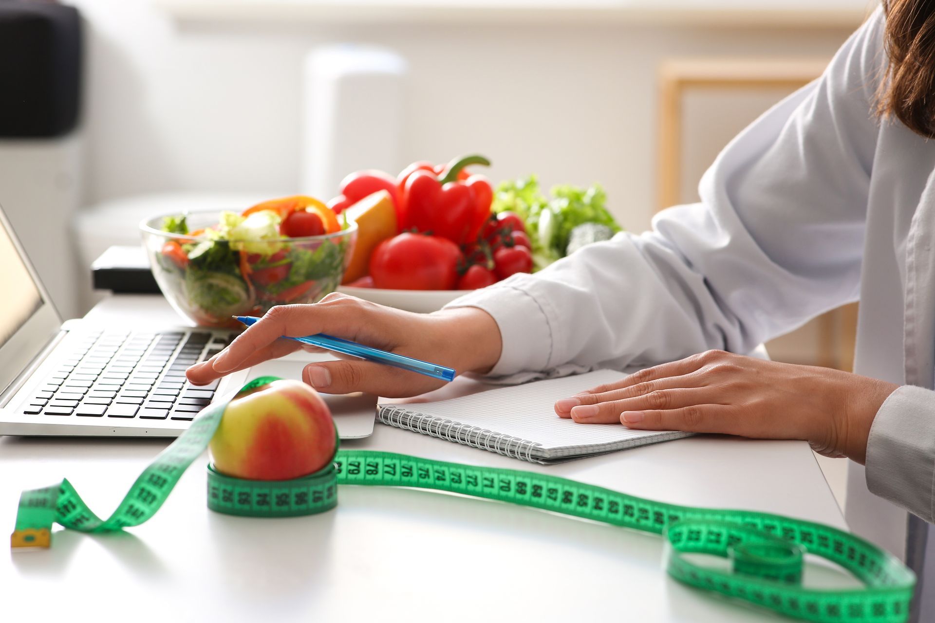 Dietician working on laptop, with notepad, measuring tape, and fruit on desk near bowl of vegetables.