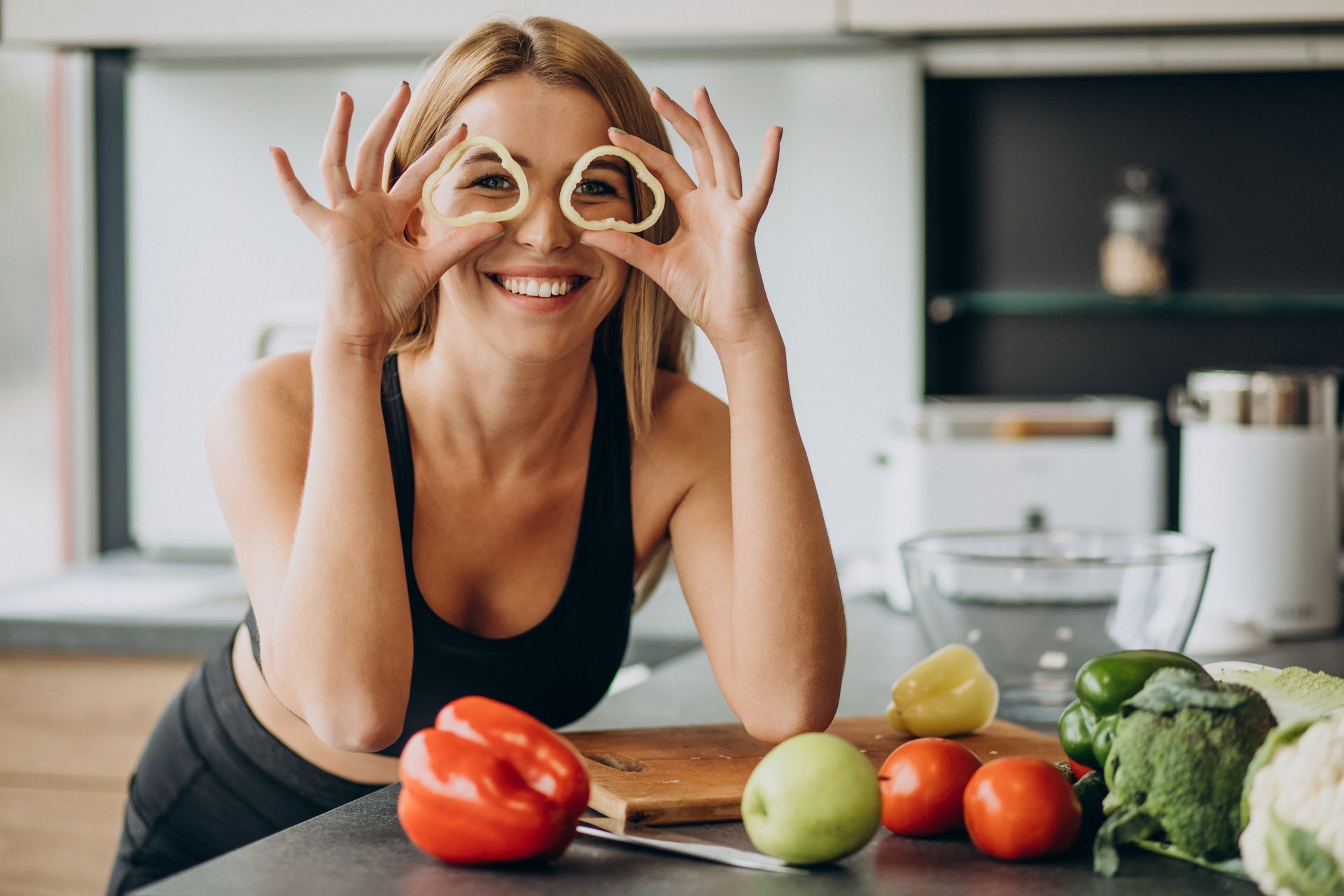Woman smiling in a kitchen, holding vegetable rings over her eyes. Vegetables on a counter.health
