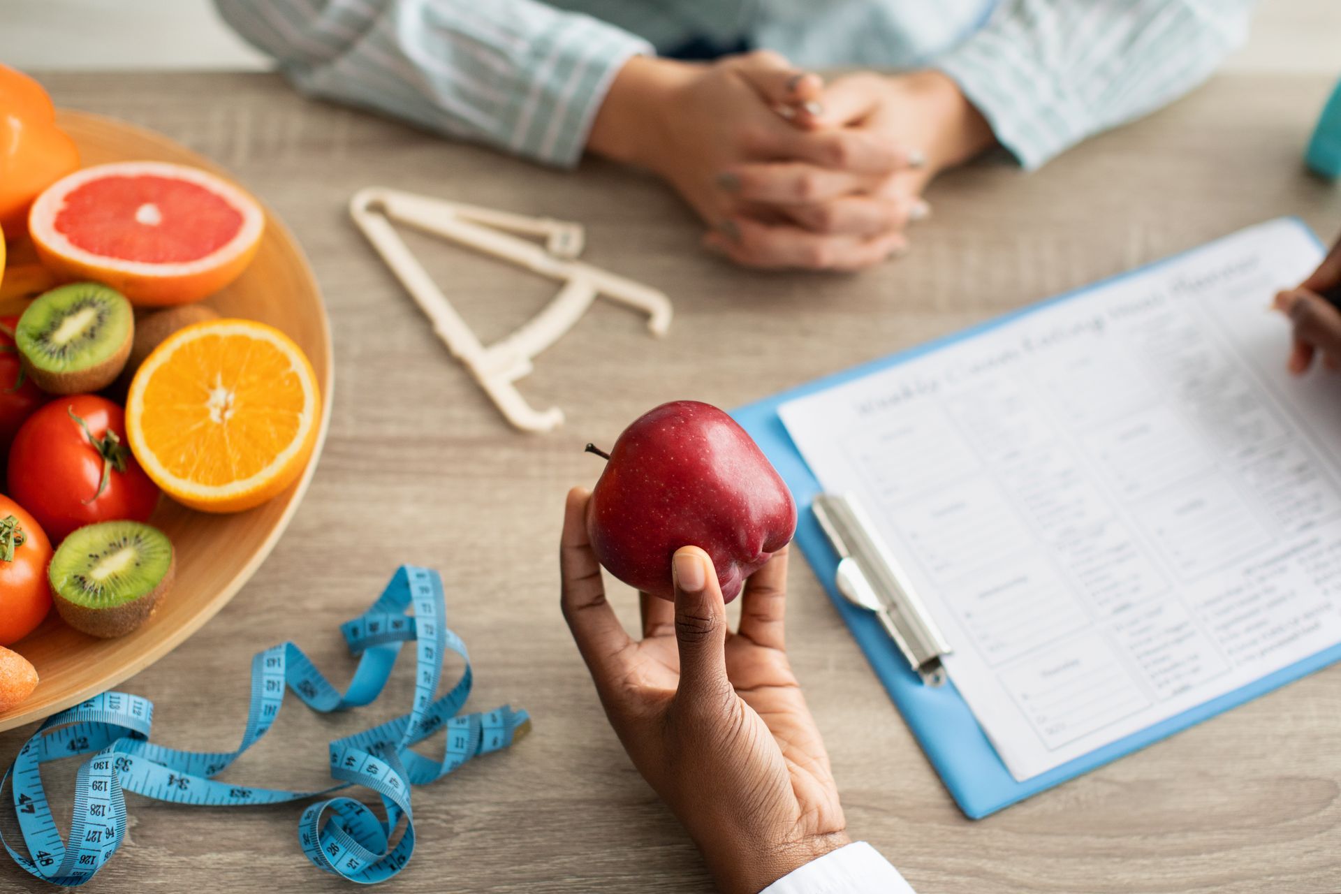 Dietician holding apple, reviewing a patient's chart, with fruit bowl and measuring tape on table.