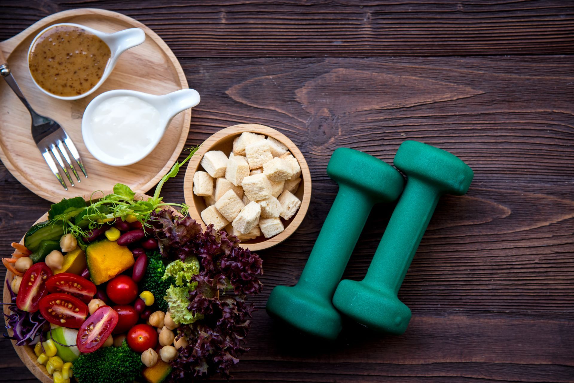 Healthy meal with tofu, dressing, and dumbbells on a wooden table.