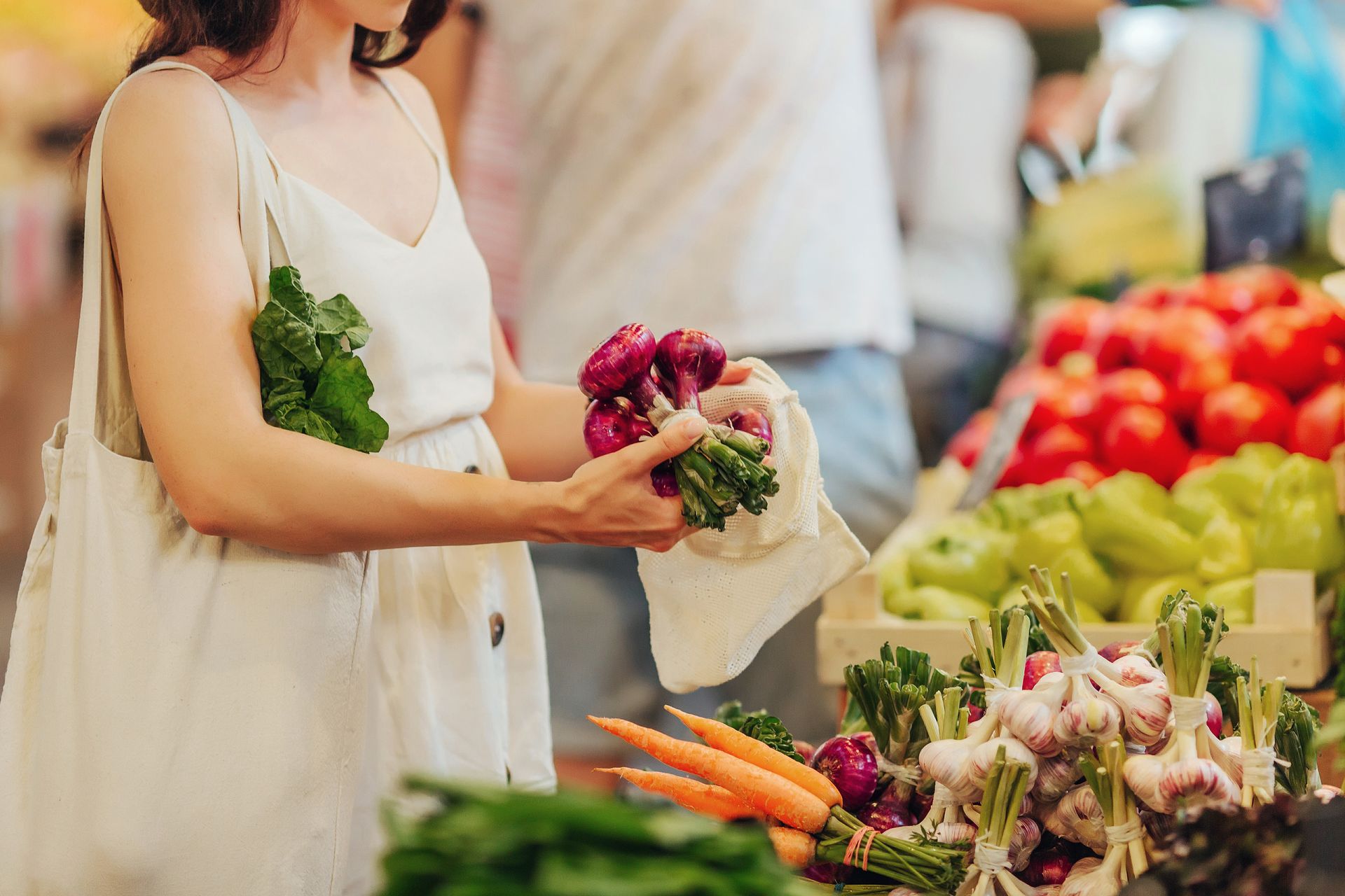 Woman shopping for produce at a market, holding red onions and reusable bag. Vegetables on display.y