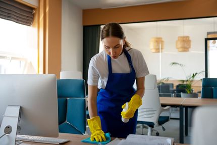 Woman in apron and gloves cleaning desk in office.