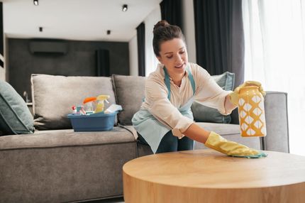 Woman cleaning a wooden coffee table in a living room, wearing gloves and an apron, with a vase.