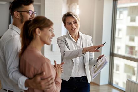 Real estate agent showing property to a couple; agent points toward a window.