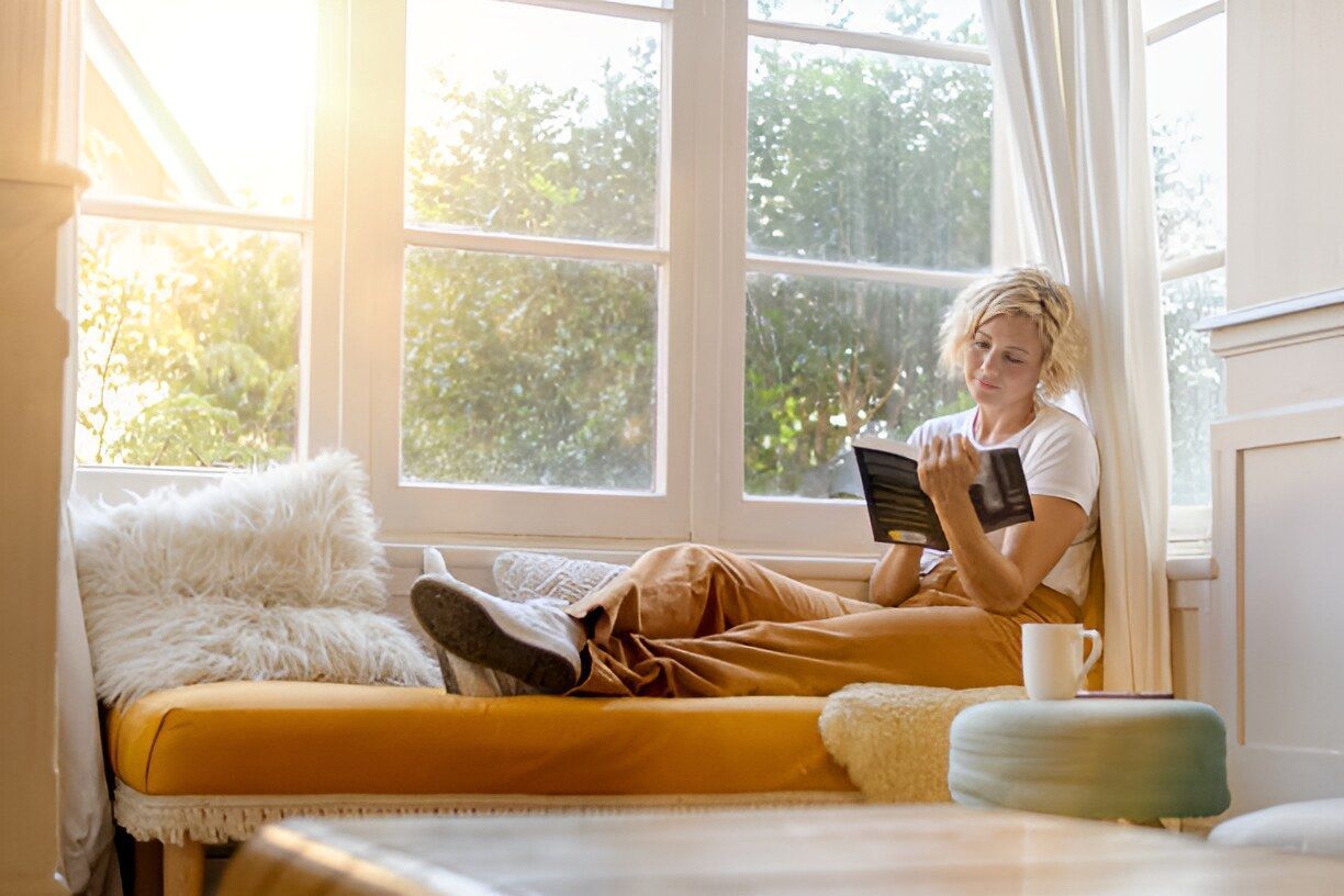 A Woman Is Sitting On A Window Sill Reading A Book — Austral Insulation In Toormina, NSW