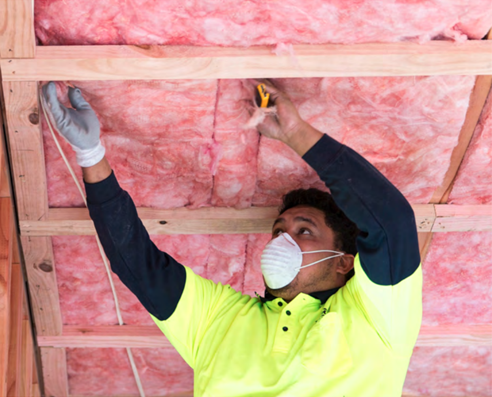 A Man Is Installing Pink Batts Insulation Into A Ceiling With A Stanley Knife — Austral Insulation In Toormina, NSW