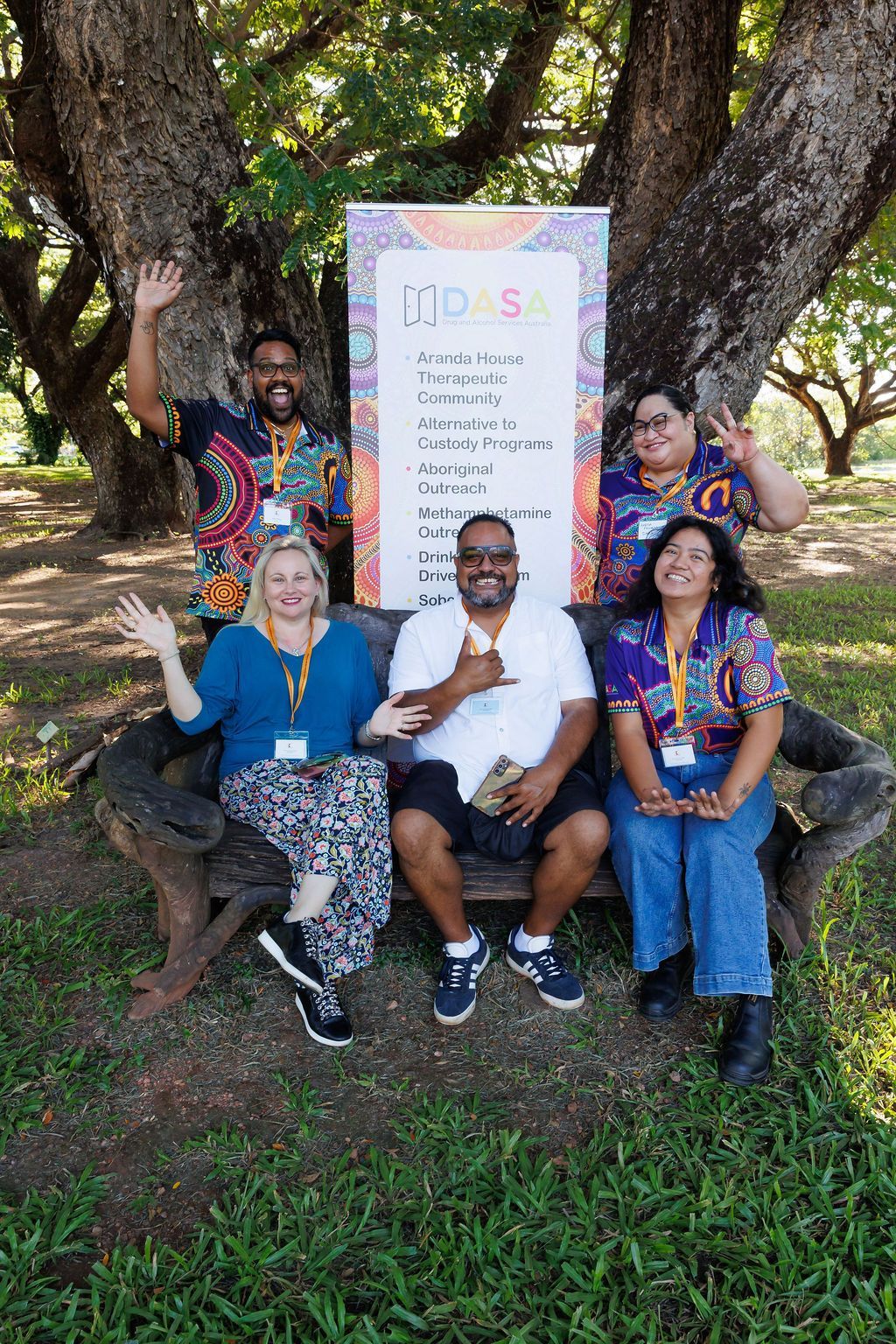 Five people pose smiling and waving on a large bench in a wooded area in front of an event sign for DAS.