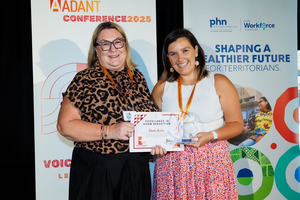 Two people standing in front of a conference banner, one holding a certificate and award while smiling for a photo.