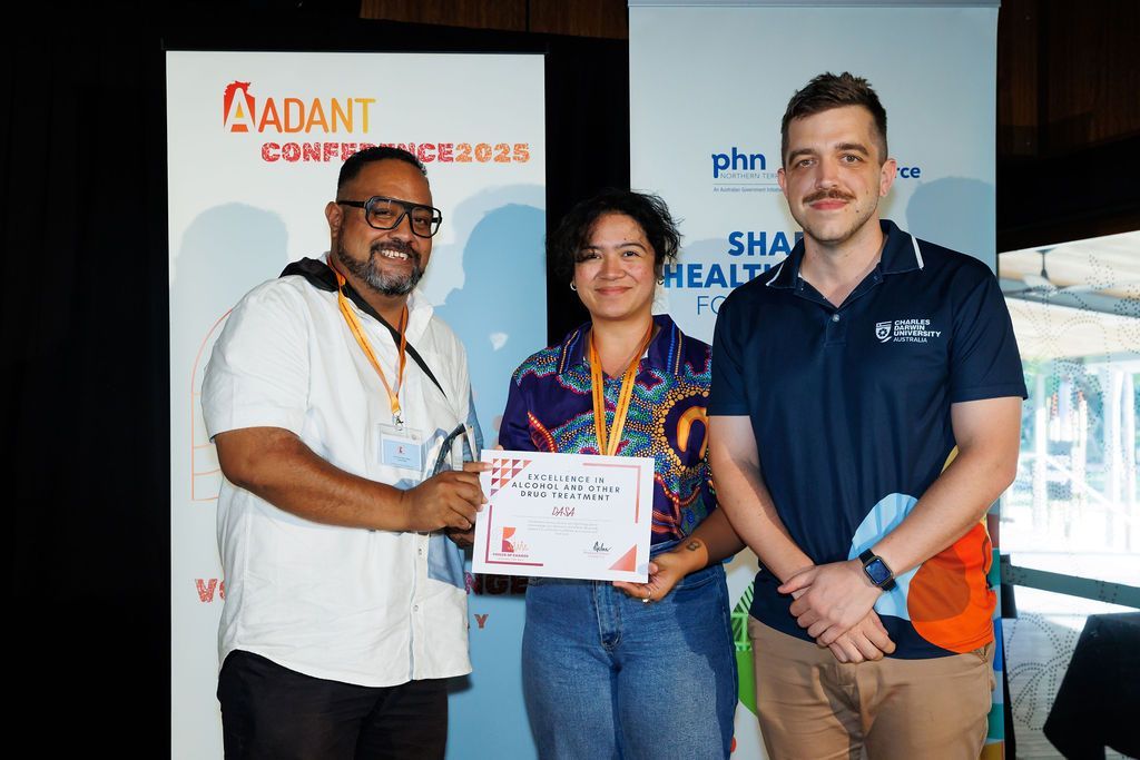 Three people stand before conference banners, holding a certificate during an event.