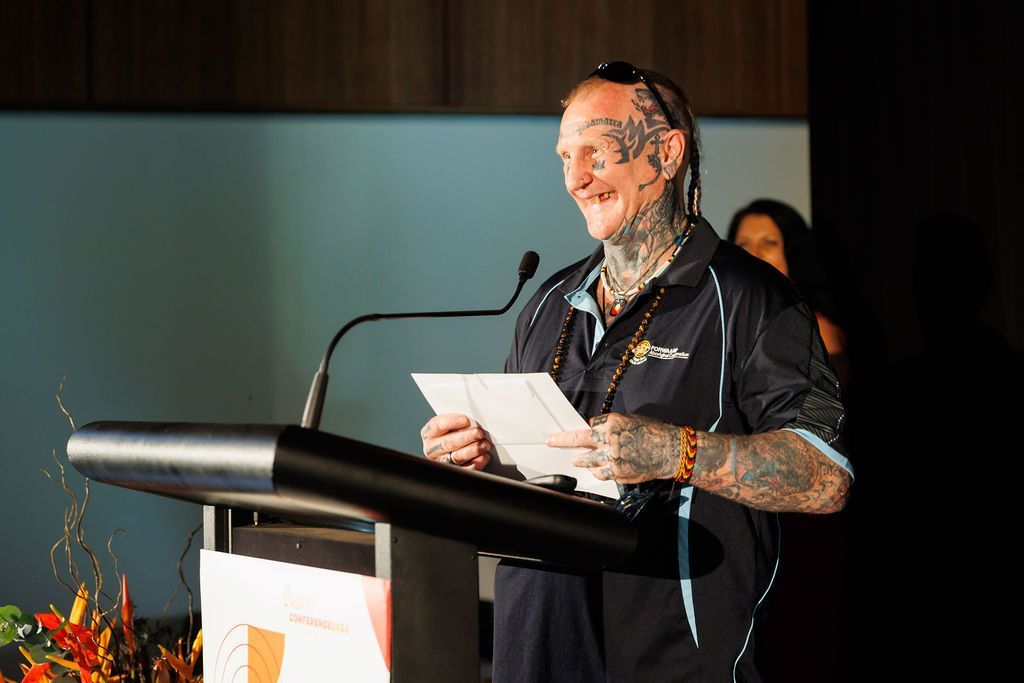 A smiling person with extensive tattoos on their face and arms speaks into a microphone at a podium while holding papers.