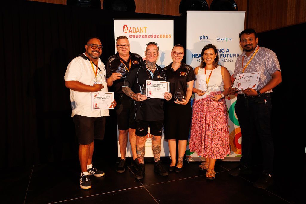 Six people stand in a line on a stage, smiling and holding certificates and awards in front of event banners.