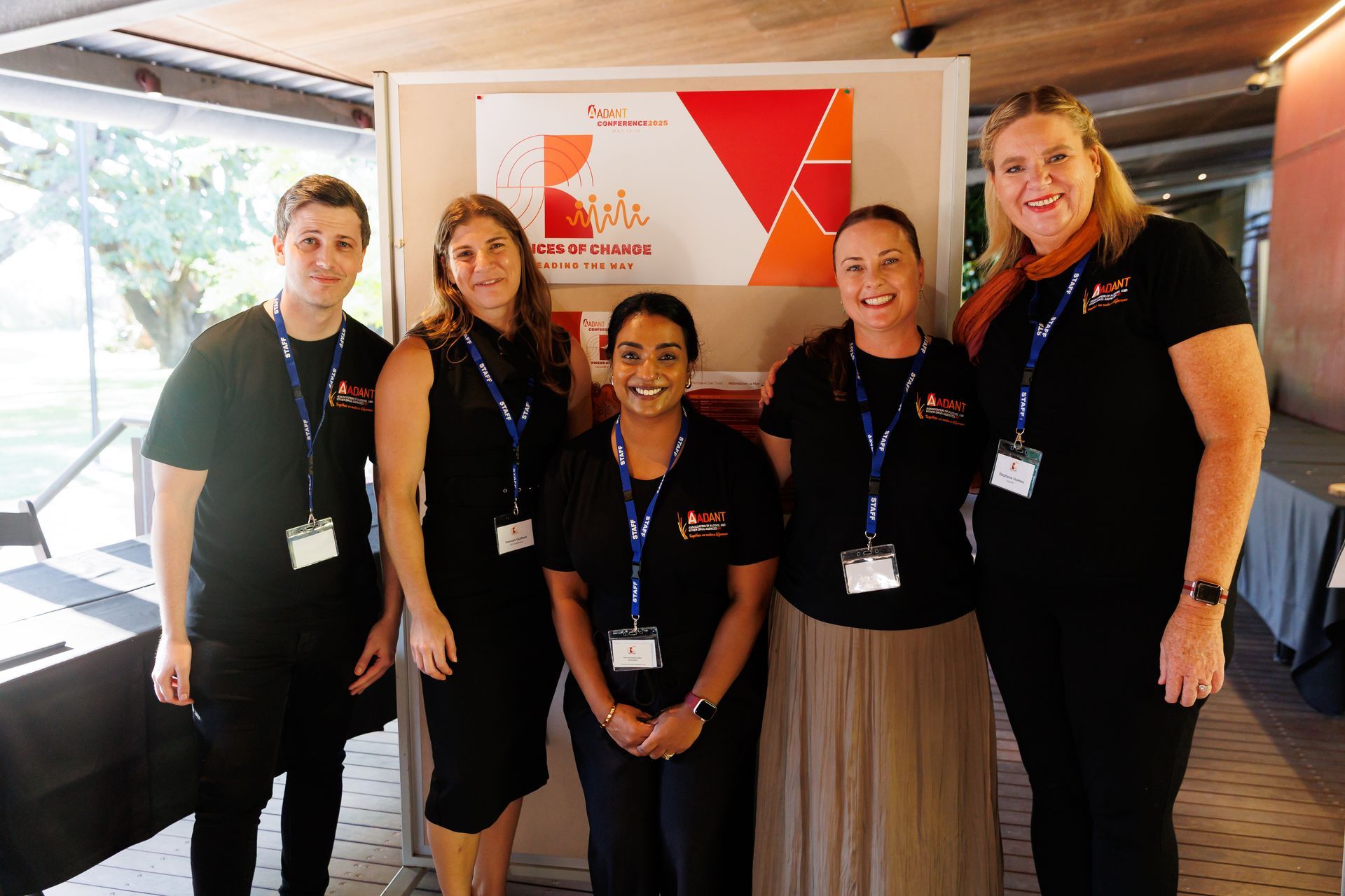 Five smiling people wearing matching black shirts and lanyards stand in a line in front of a conference event banner.