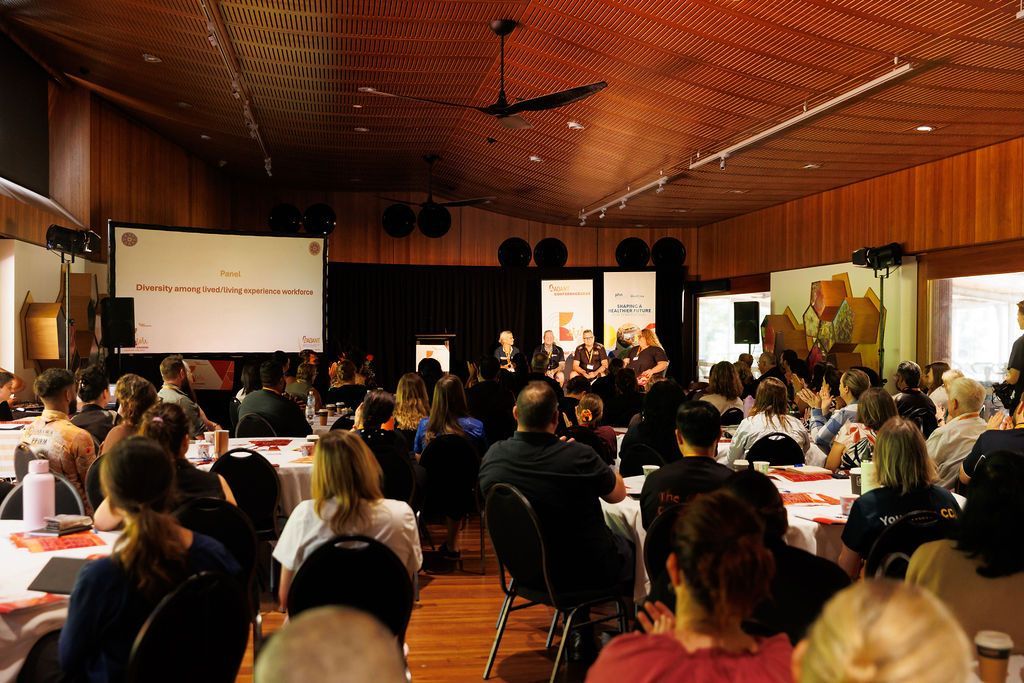 An audience seated at round tables in a wood-paneled hall, facing a presentation screen and speakers on a stage.