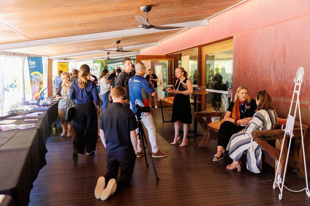 People mingle on a wooden deck during an event, with one person kneeling to film and two people seated talking.