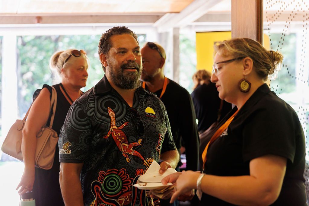 A man and woman in an indoor setting converse while holding small plates of food, with others in the background.