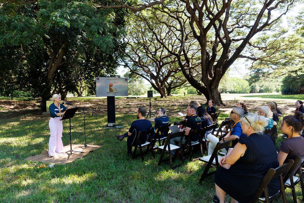 A person speaks at a podium to an outdoor audience seated in chairs under large, shady trees.