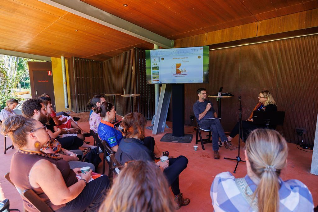 A speaker sits on stage in a covered outdoor area, addressing an audience seated in chairs facing them and a screen.