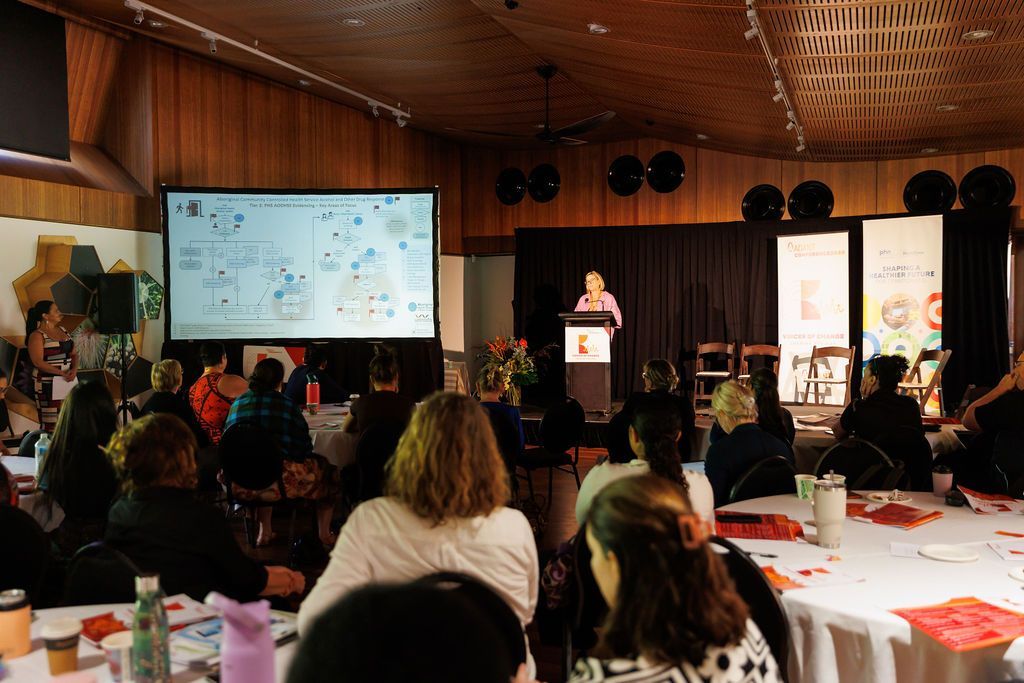A presenter speaks at a podium in a dim conference room before an audience, with a large slide projected on the screen.