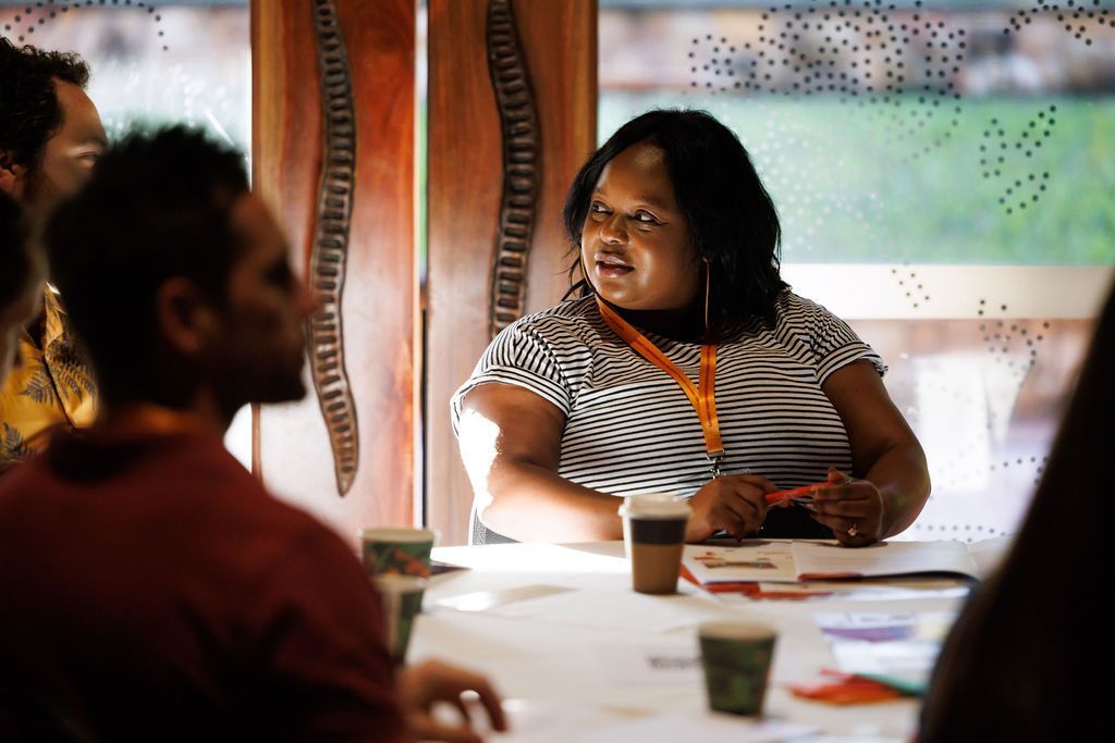 A person with an orange lanyard speaks at a table with coffee cups, facing others in a brightly lit indoor space.