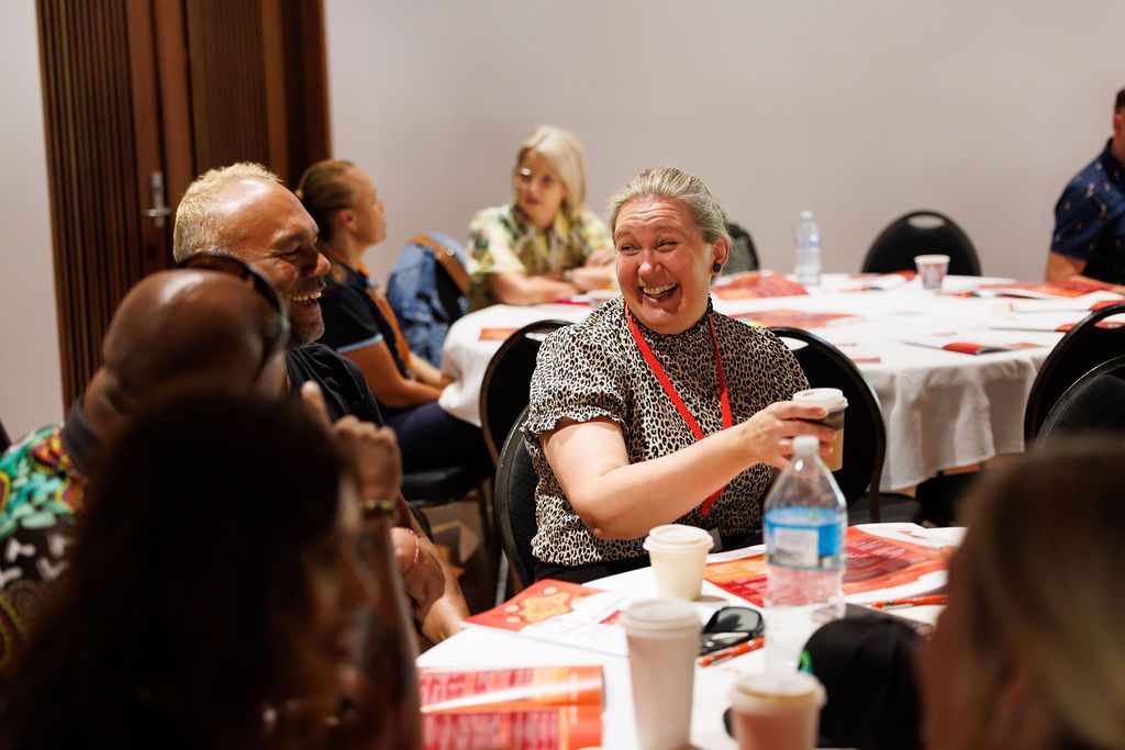 A group of people sit around tables in a brightly lit room, laughing and talking during a social gathering.