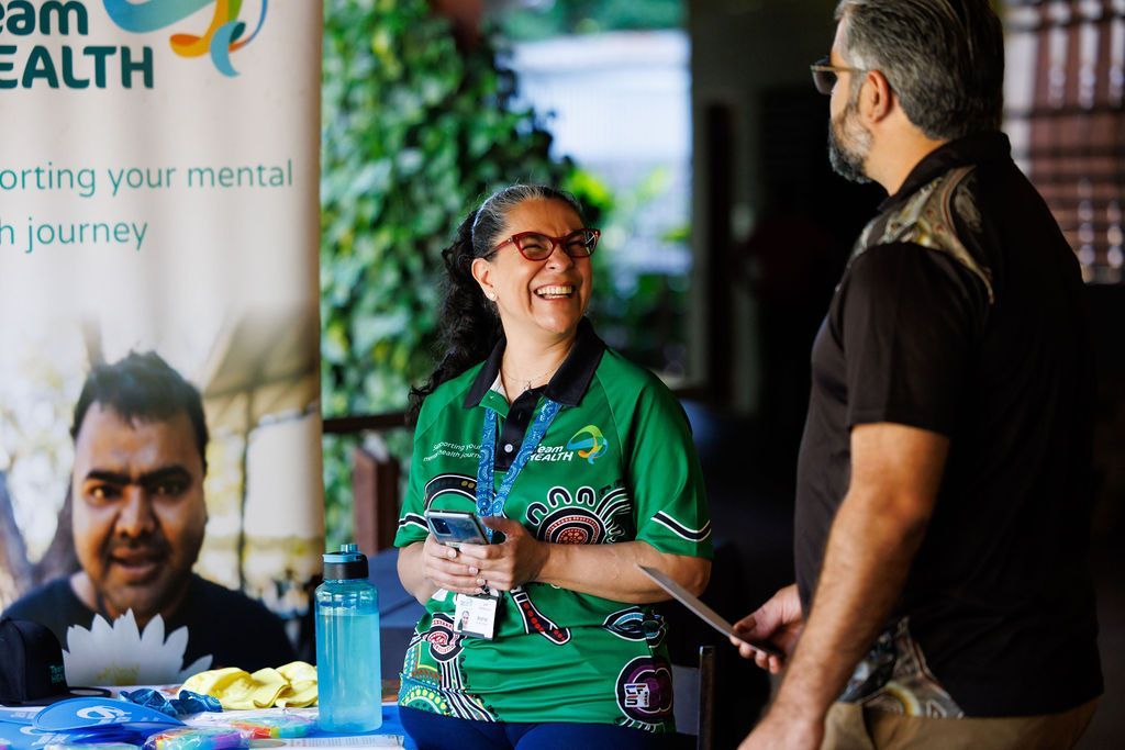 A person in a green shirt smiles while talking to another person next to a mental health awareness banner.