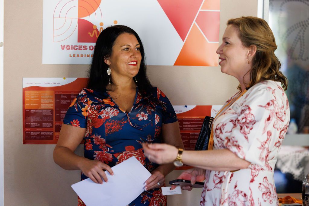 Two women stand in front of a “Voices” banner, holding documents and speaking to one another in an office-like setting.