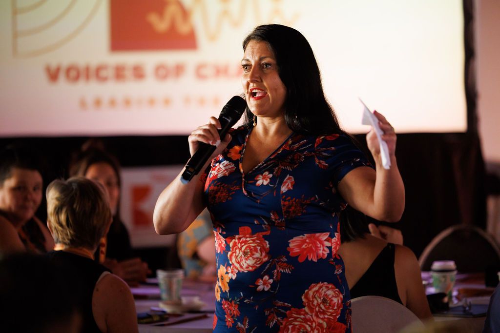 A person with dark hair in a floral dress speaks into a microphone before an audience at a 