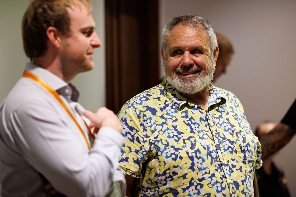Two men conversing in a brightly lit room; one wears a light shirt with an orange lanyard, the other a yellow camo shirt.