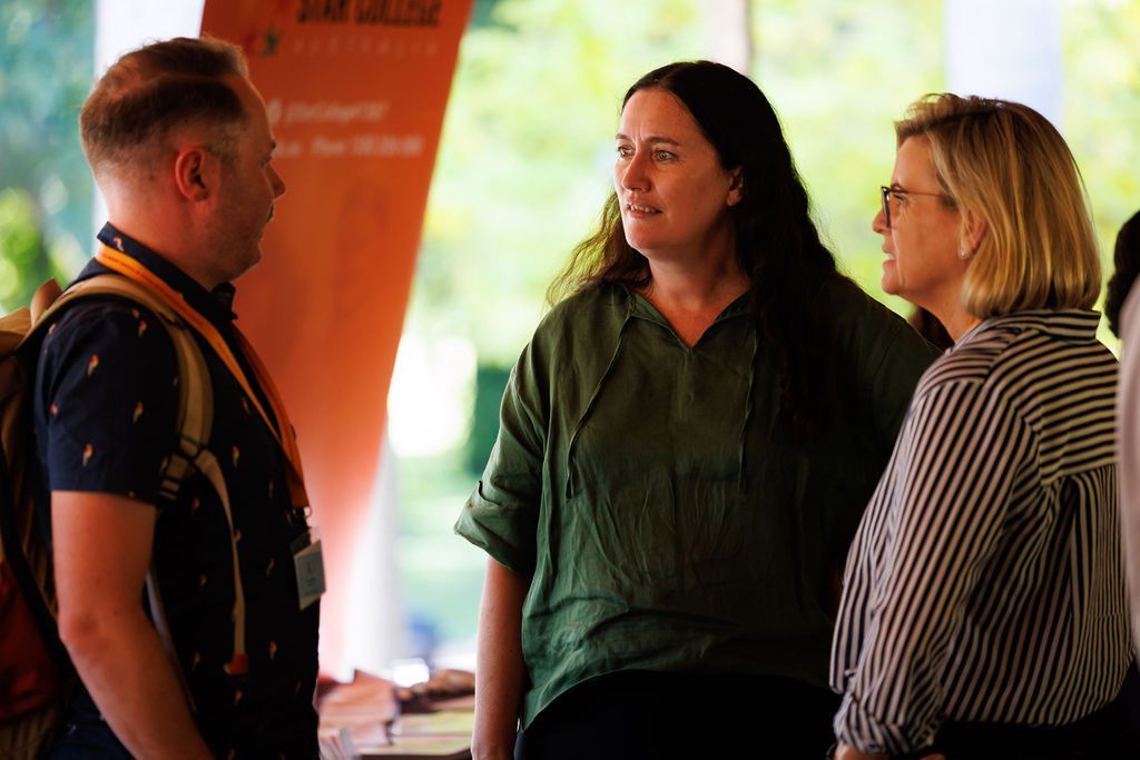 Three people engaged in a professional conversation at an indoor event, standing near an orange display banner.