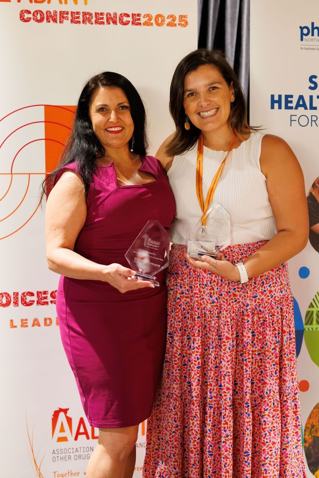 Two smiling individuals hold glass awards in front of a conference banner with orange and blue text and graphics.