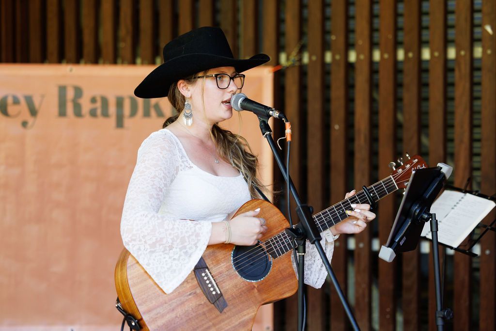 A musician in a black cowboy hat and white lace top performs while playing an acoustic guitar into a microphone.